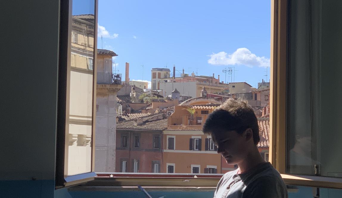 A student at the Rome Center works on their assignment with the window open. Outside the window are terracotta-colored buildings.