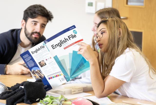 Three students in English class, one holding book that two are reading. 