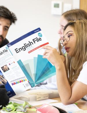 Three students in English class, one holding book that two are reading. 