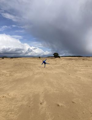 A student stands on a large patch of dirt with large clouds looming in the background. The student is standing on one foot in such a way that they look like a starfish.