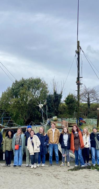 large group of students standing on a sand beach in front of a massive boat with grass, trees, and fog in the background