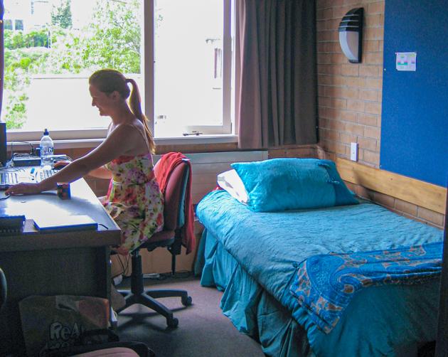A student works at their desk in a small bedroom with a large window, a bed, and comfortable seating.