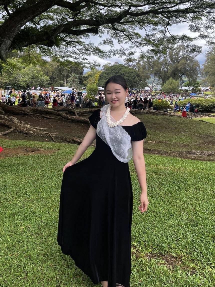 木の下で、黒いハワイアンワンピースと首元に白いレイを身に着けた姿